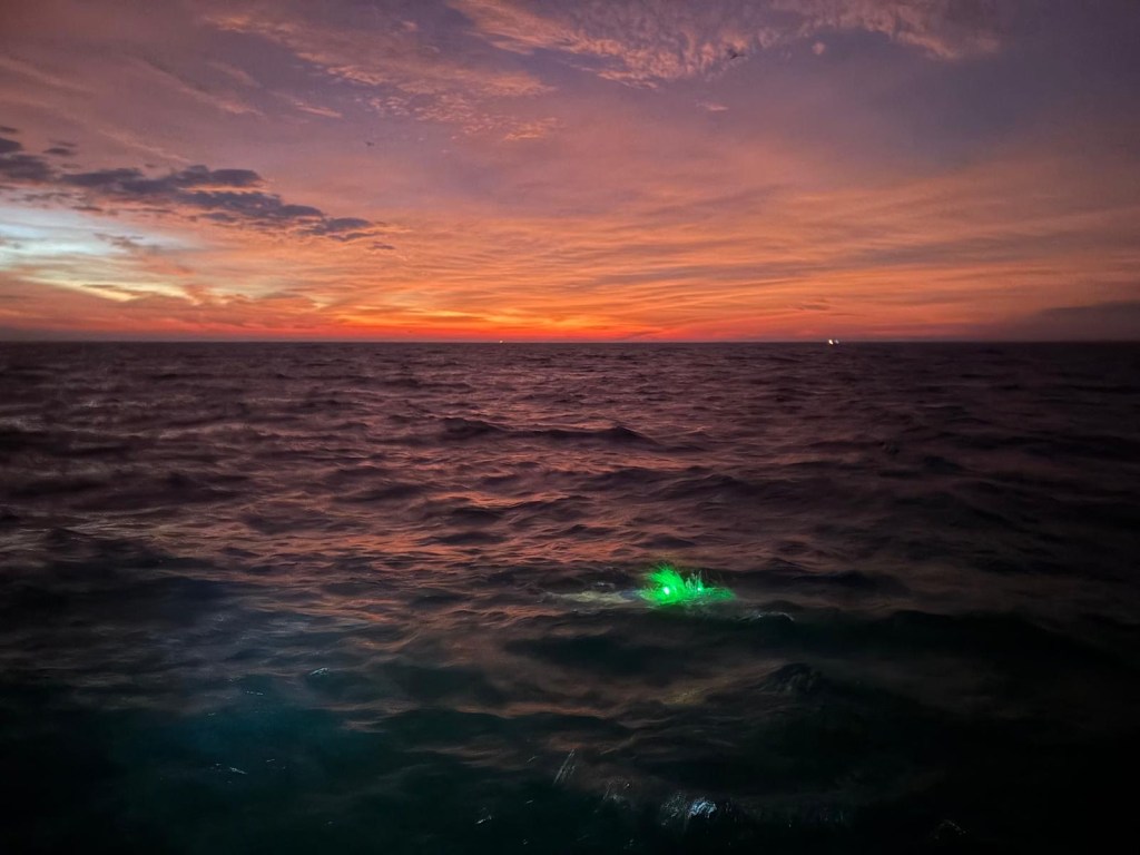 A serene ocean view at dusk, featuring a colorful sunset over the water, with gentle waves and a glowing green light visible in the foreground.