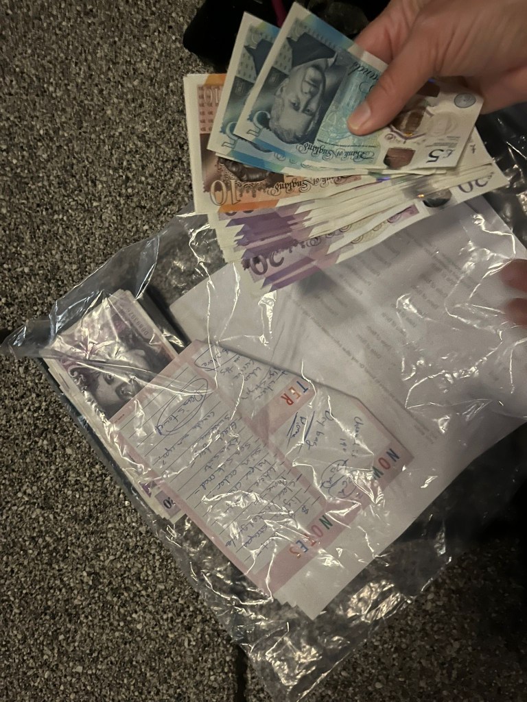A hand holding various banknotes while other notes and documents are visible inside a clear plastic bag on a textured surface.