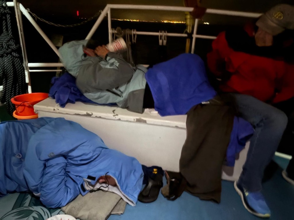 Two individuals resting on a boat at night, one sitting on a cooler wearing a red jacket and a cap, while another is lying down wrapped in blue blankets.