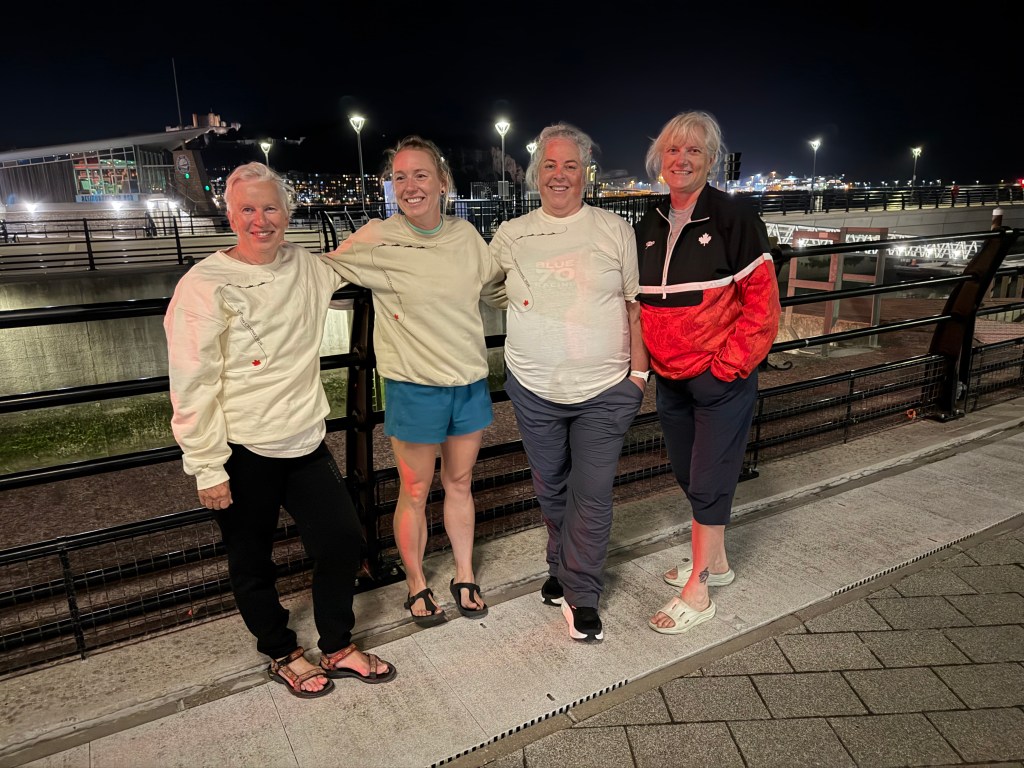Four women posing for a photo at night near a waterfront, wearing casual clothing including sweatshirts and shorts, with city lights in the background.