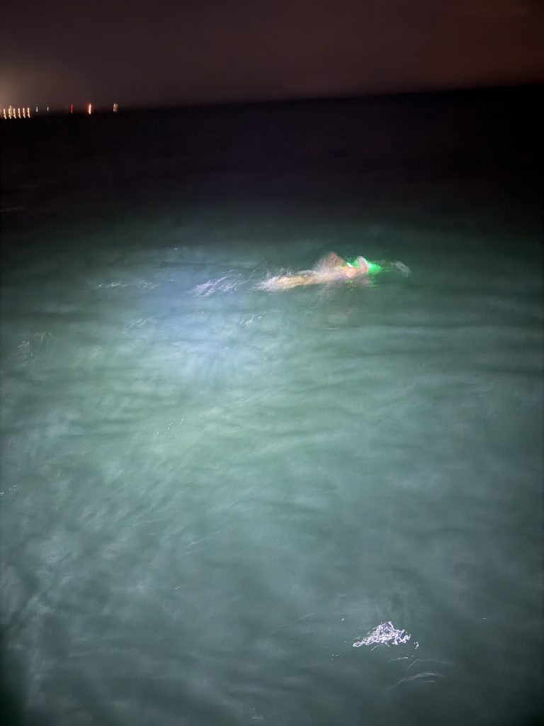 A person swimming in dark waters at night, illuminated by a light source, with faint city lights visible in the background.