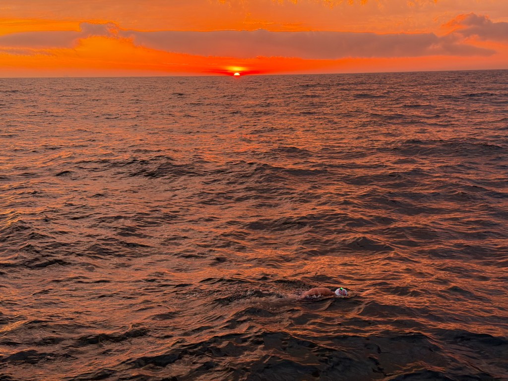 A swimmer wearing a colorful cap glides through ocean waters at sunset, with vibrant orange and yellow hues lighting up the sky.