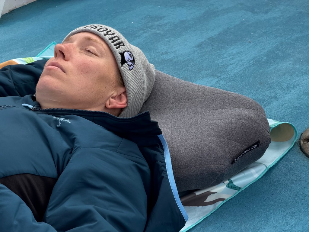 A person resting on a blue surface, wearing a winter hat and a jacket, with a large gray pillow positioned behind their head.