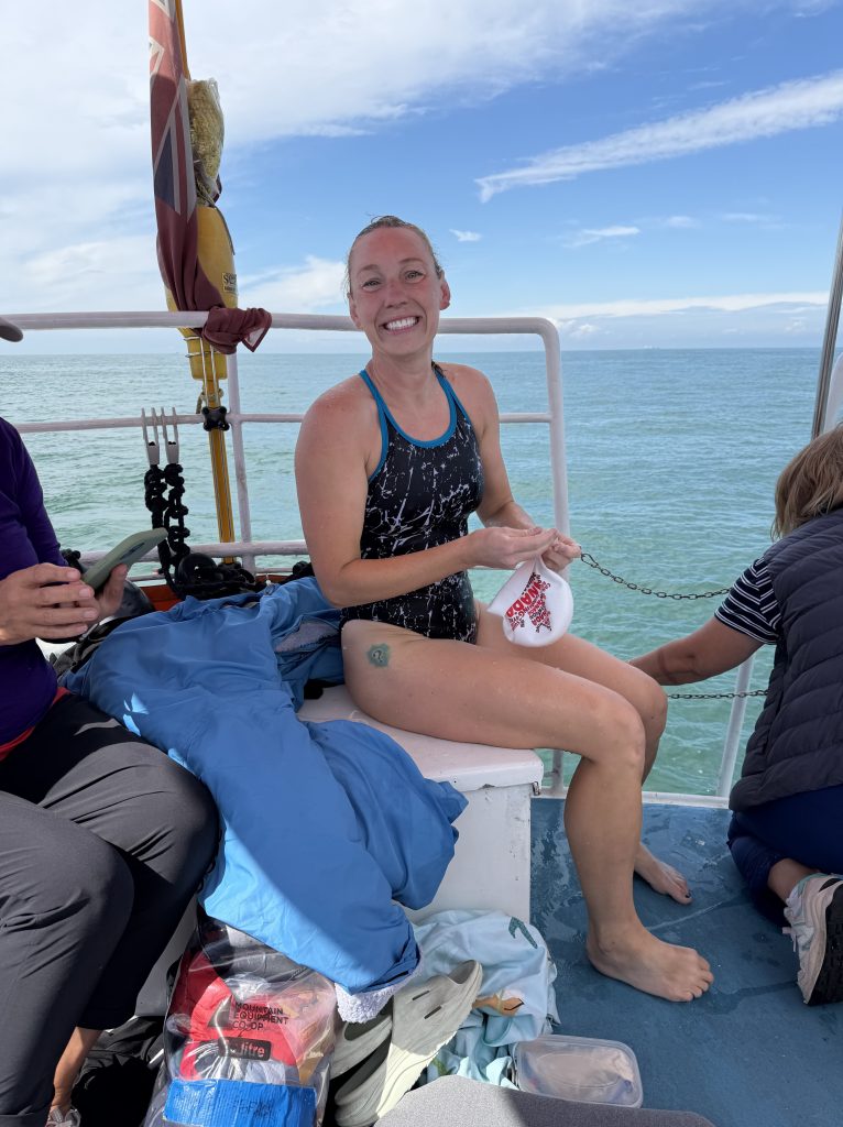 A smiling woman in swimwear sitting on a boat, holding a plastic bag, with people and bags in the background, overlooking calm waters under a blue sky.