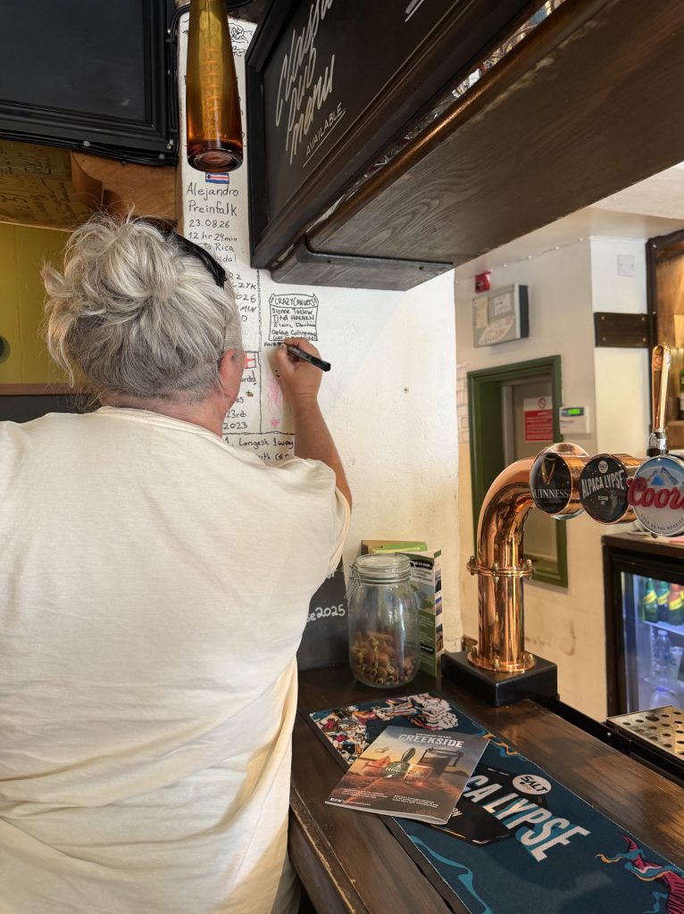 A person with light gray hair, wearing a white shirt, is writing on a wall in a pub. Behind them is a bar with beer taps and a menu displayed on the counter.