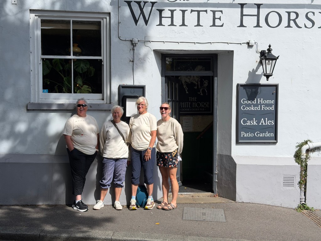 Four women standing outside a pub called 'The White Horse,' smiling and wearing matching light-colored shirts. The pub features a sign with details about food and beverages.