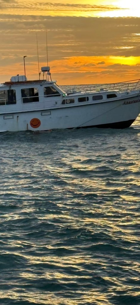 A boat named 'Anastasia' sailing on shimmering water during sunset, with waves reflecting shades of orange and gold in the background.